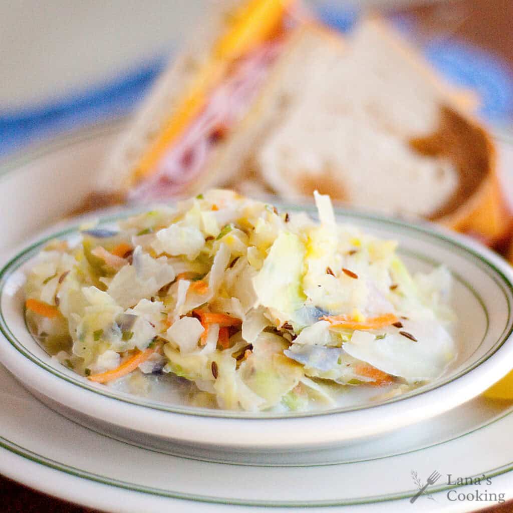 A bowl of creamy hot cabbage slaw with a sandwich on a plate in the background.
