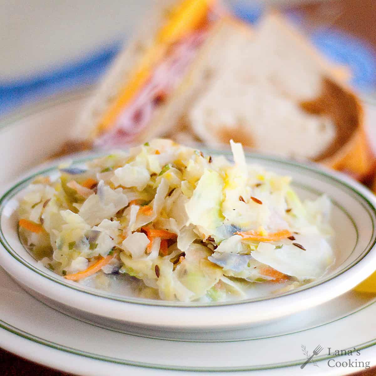 A bowl of creamy hot cabbage slaw with a sandwich on a plate in the background.
