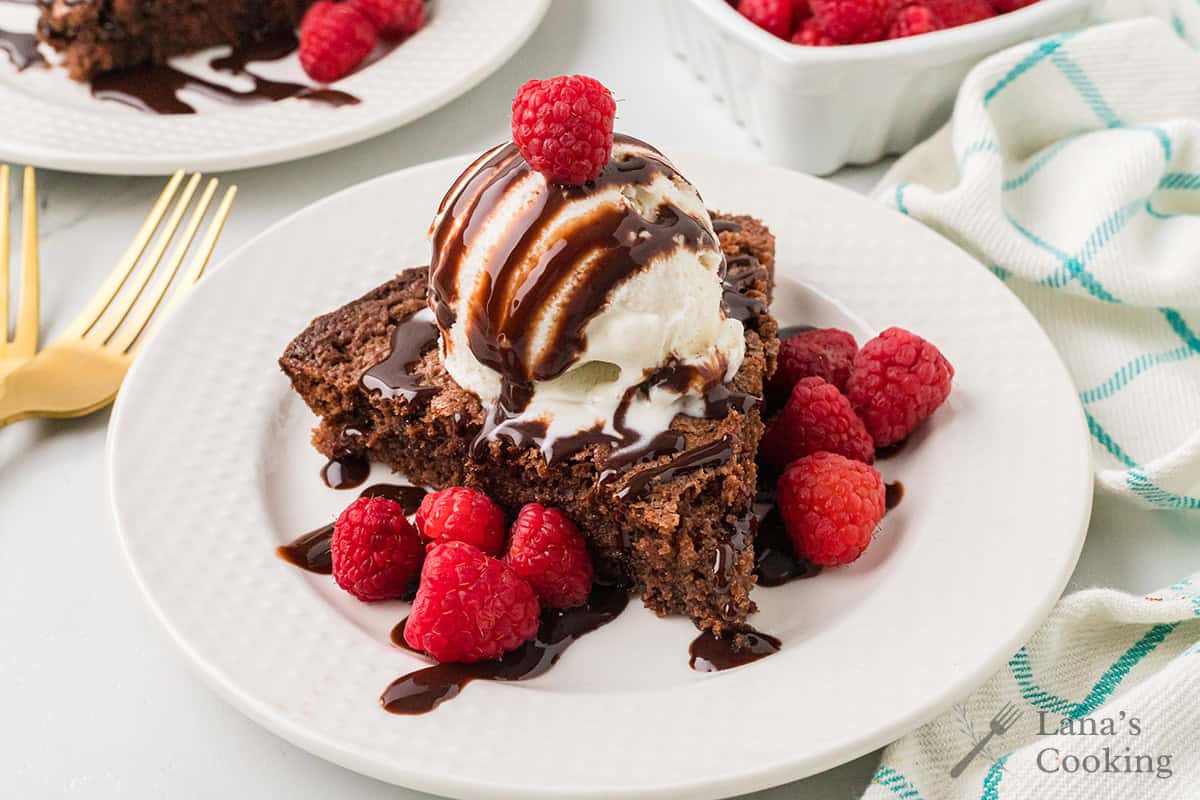 A slice of hot fudge pie on a white plate with a drizzle of chocolate syrup and vanilla ice cream surrounded by fresh raspberries.