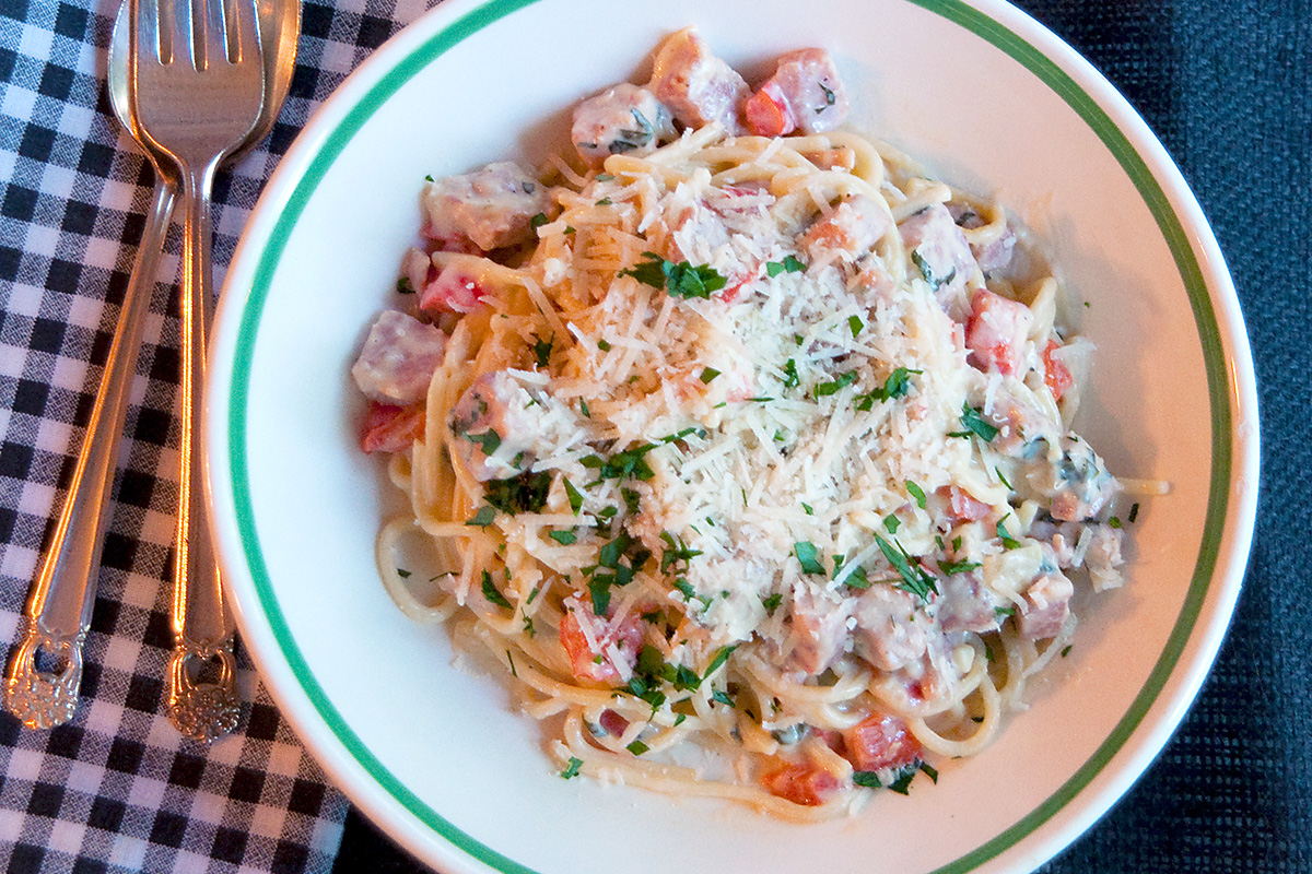 A bowl of pasta topped with a creamy sauce,, herbs, and grated cheese, with a fork and spoon on a checkered napkin next to the plate.