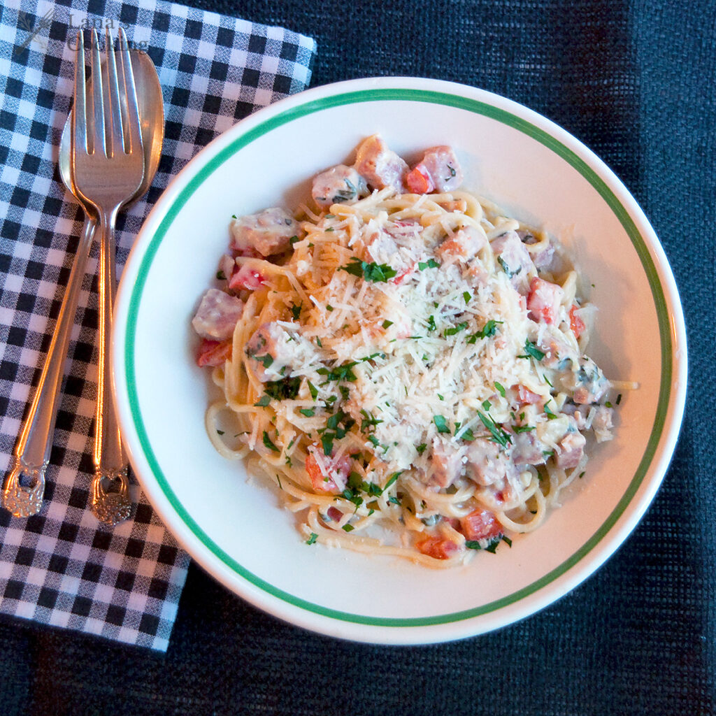 A bowl of pasta with creamy sauce, diced vegetables, and grated cheese, placed on a gingham napkin with a fork and spoon beside it.