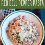 A plate of creamy ham and red bell pepper pasta topped with grated cheese and herbs, placed on a white plate with a fork and knife beside it.