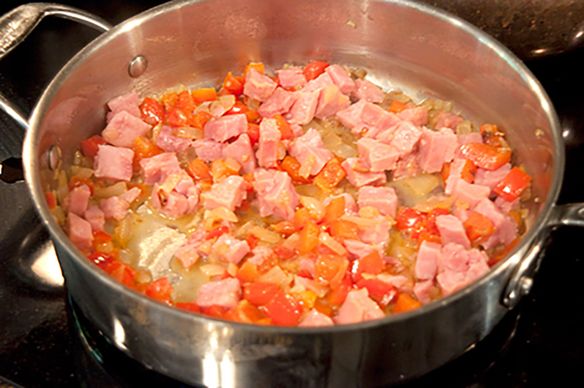 Diced ham and chopped red bell peppers cooking in a stainless steel sauté pan on a stovetop.