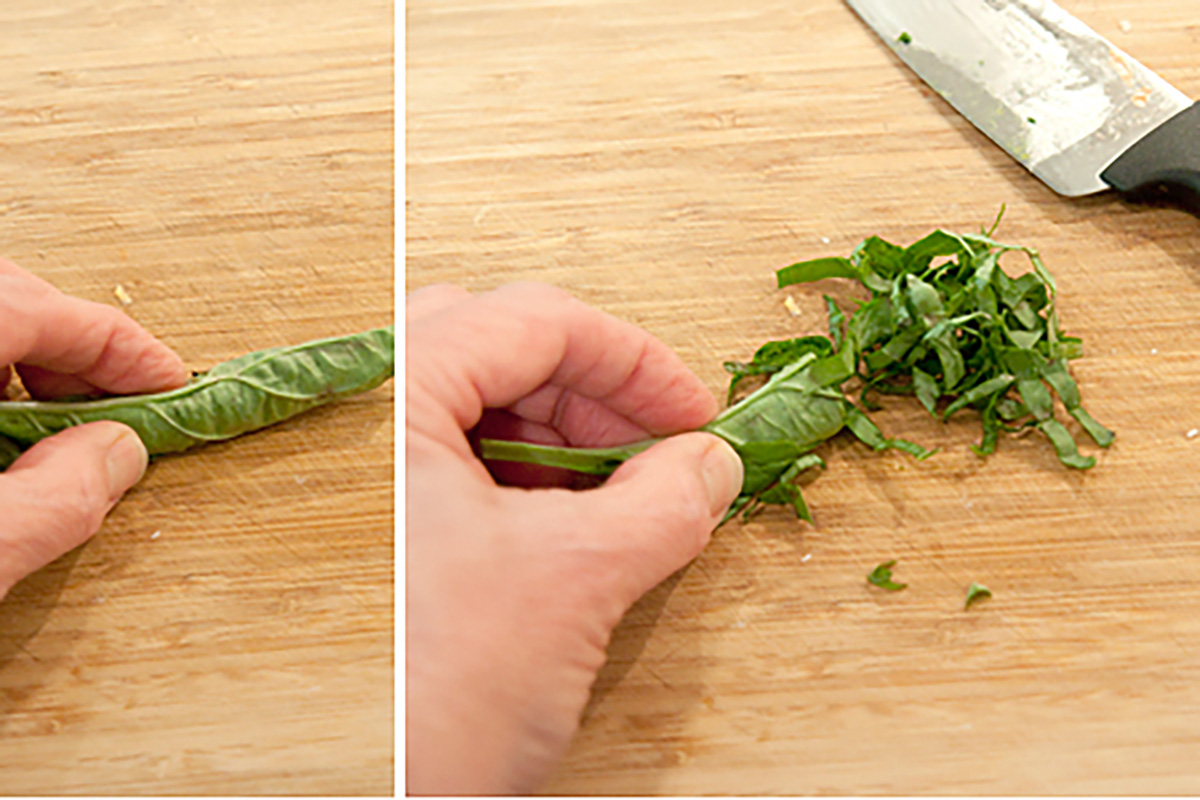 A hand rolls basil leaves, then slices them into thin strips on a wooden cutting board with a knife nearby.