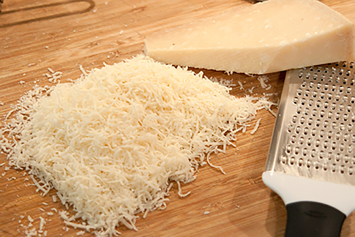 A pile of grated cheese next to a wedge of cheese and a cheese grater on a wooden cutting board.