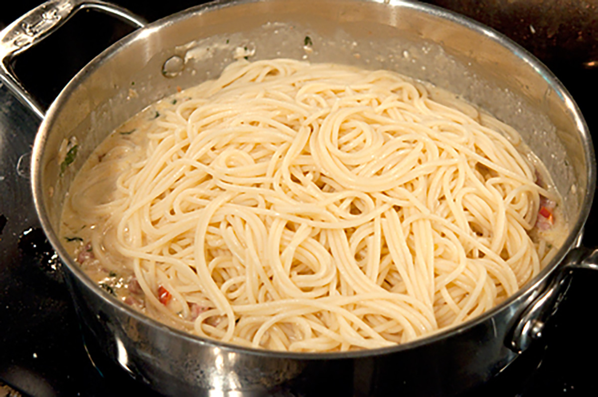 A stainless steel pan on a stovetop filled with cooked spaghetti noodles in a creamy white sauce.