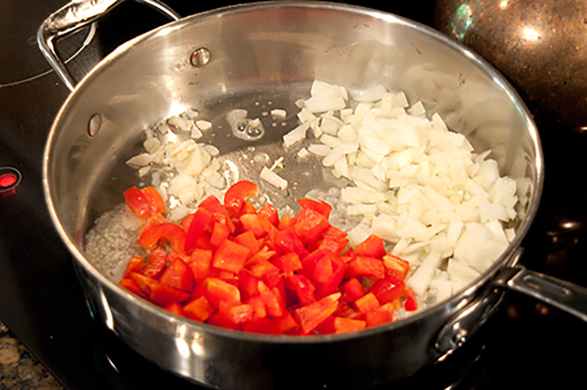 Chopped onions and red bell peppers cooking in a stainless steel pan on a stovetop.