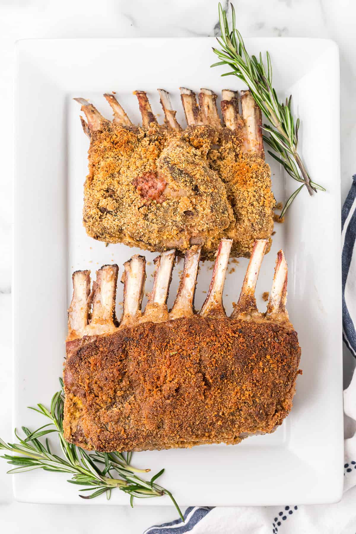 Two racks of herb-crusted lamb on a white platter, garnished with fresh rosemary sprigs.