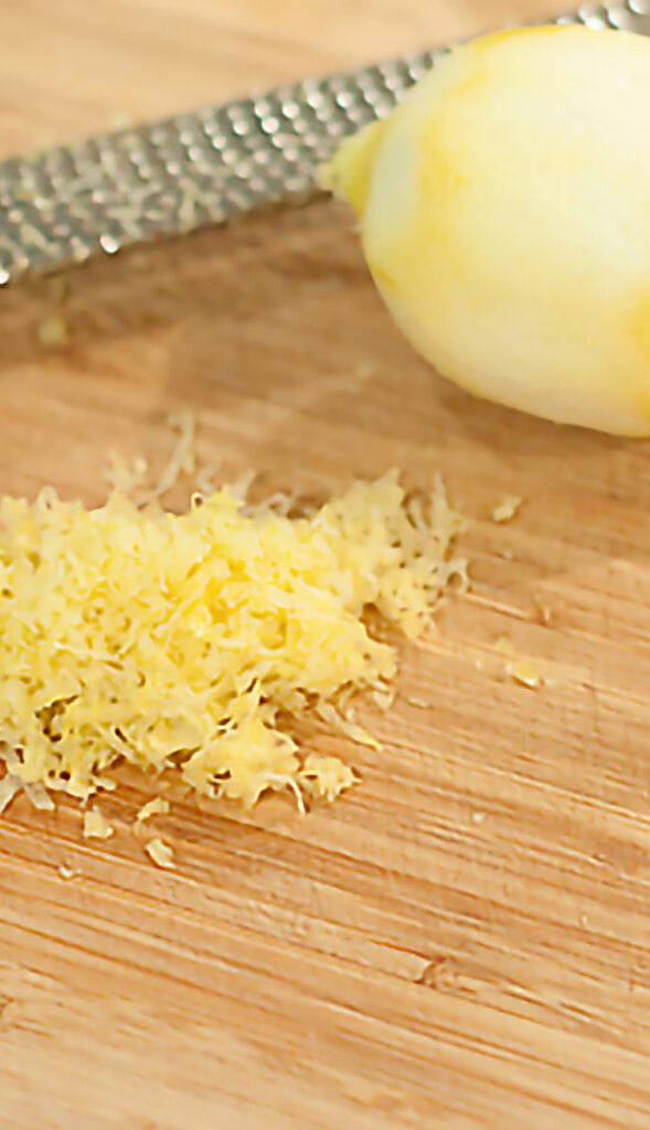 A zested lemon, a pile of lemon zest, and a grater on a wooden cutting board.