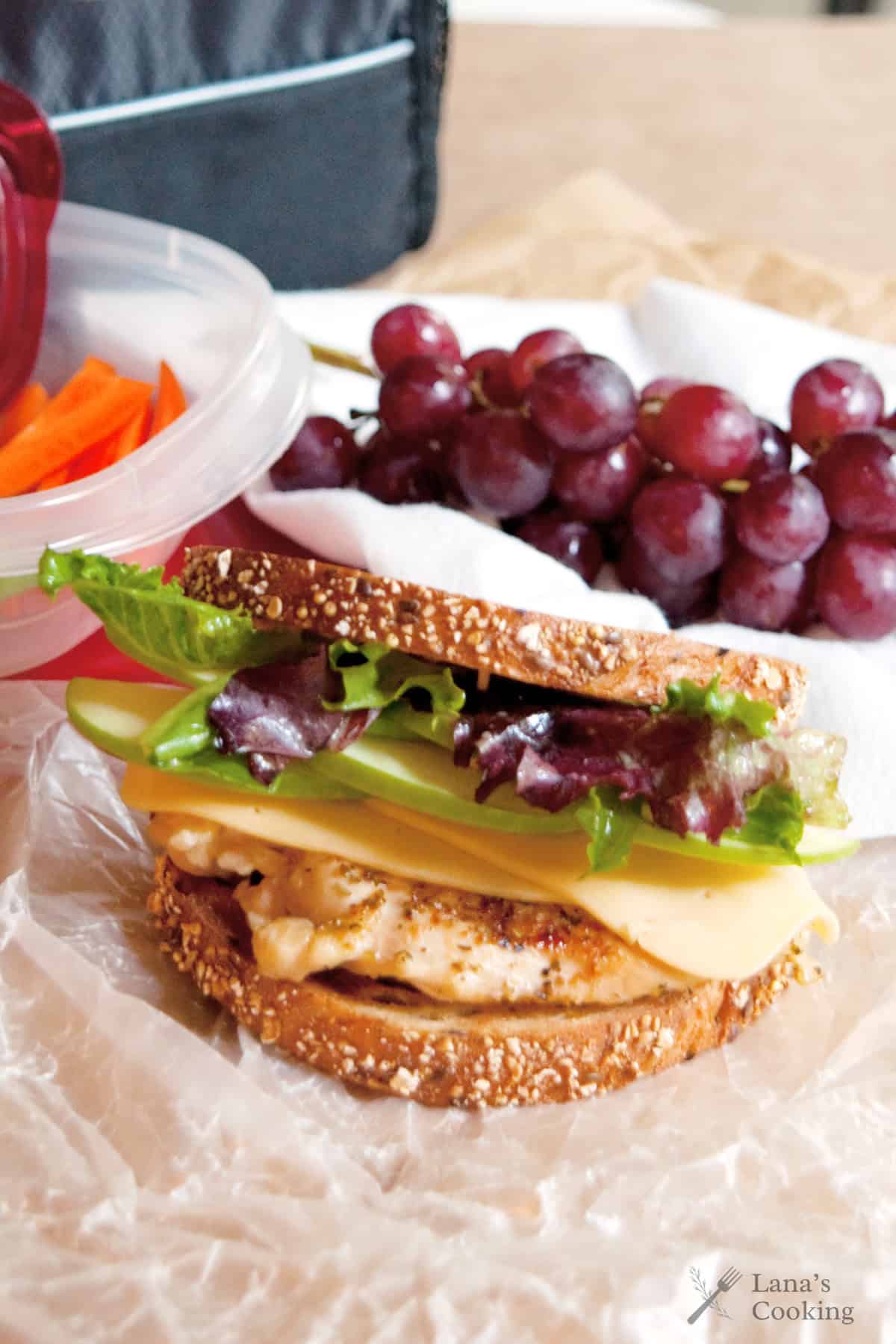 A sandwich with leafy greens, cheese, apple slices, and grilled chicken on multigrain bread sits on wax paper. Behind it are a container of carrot sticks and a bunch of red grapes.