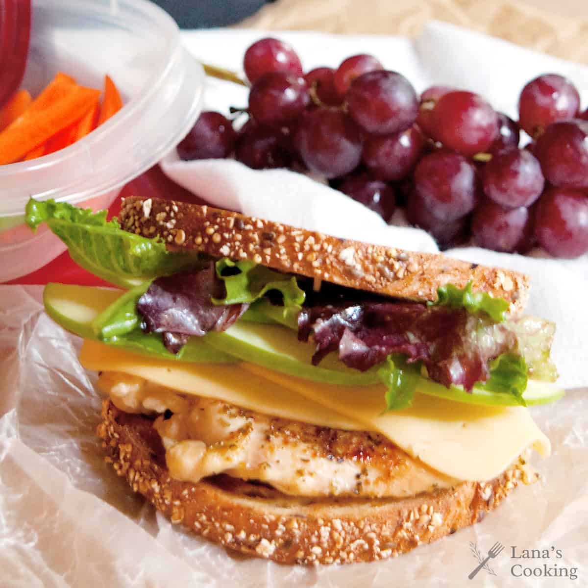 A sandwich with lettuce, cheese, chicken, and whole grain bread sits on wax paper. A bunch of red grapes and a plastic container with carrot sticks are nearby. The image includes a watermark that reads Lanas Cooking.