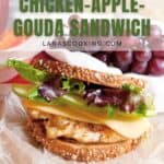 A chicken, apple, and Gouda cheese sandwich with lettuce on multigrain bread sits on wax paper, with grapes and a red container in the background. Text reads Workday Lunch! Chicken-Apple-Gouda Sandwich.
