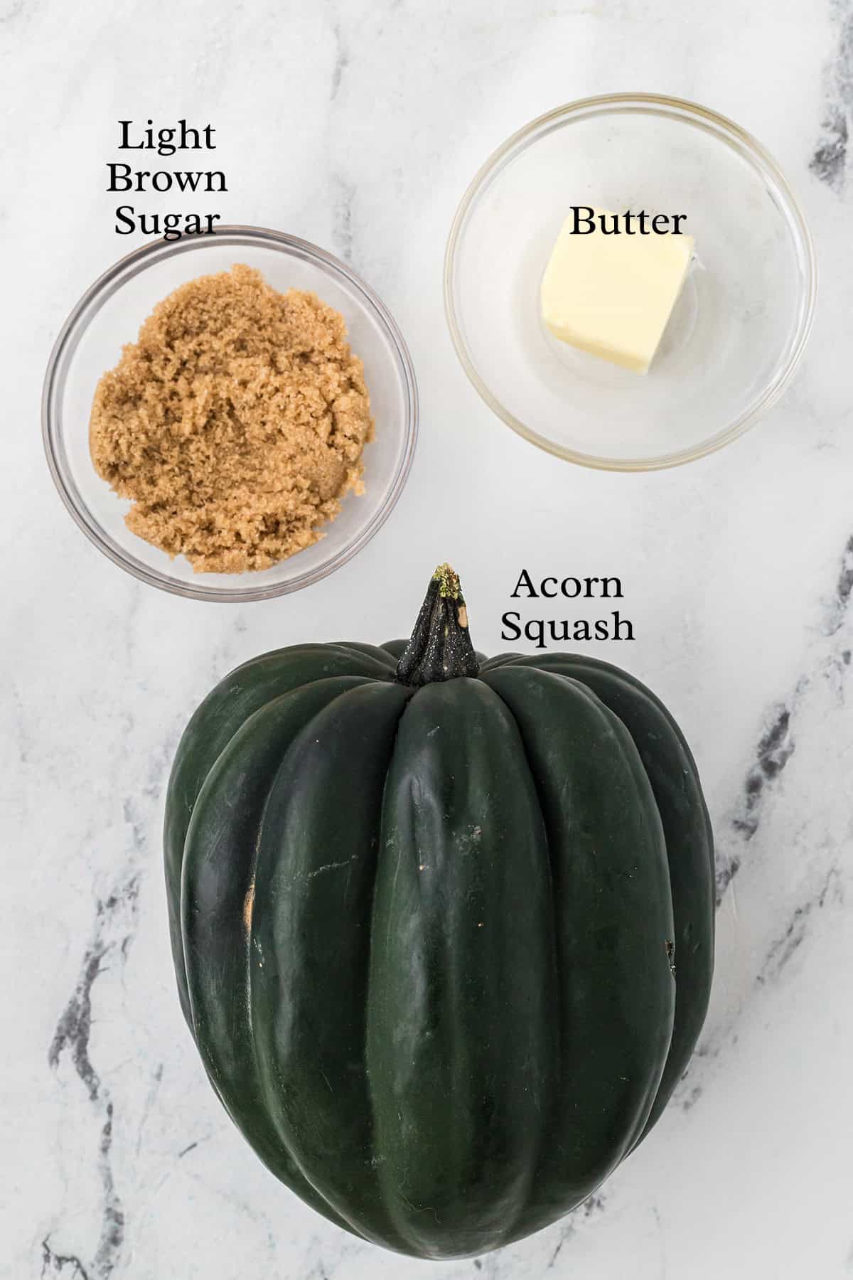 Acorn squash, a bowl of light brown sugar, and a dish of butter on a marble surface, labeled.