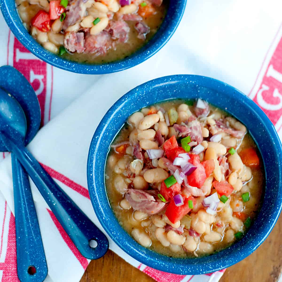 Two bowls of Slow Cooker White Beans with Smoked Ham Hocks presented on a wooden board with a kitchen towel and spoons.
