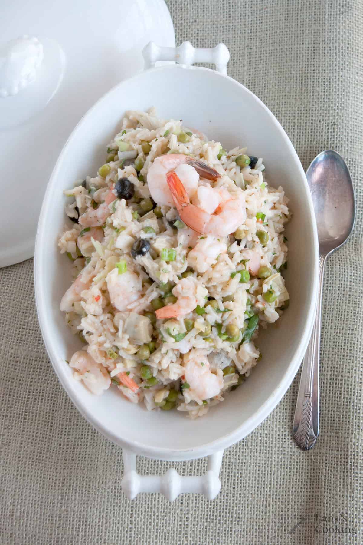 White oval dish of rice with shrimp, vegetables, and a spoon on a textured beige tablecloth.