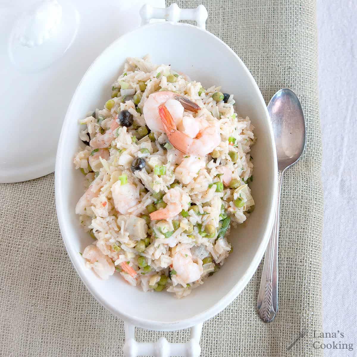 Oval dish of shrimp and rice salad with vegetables, placed next to a spoon on a light-colored cloth.