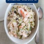 Oval bowl of shrimp and rice salad with peas and vegetables, next to a spoon on a textured tablecloth.
