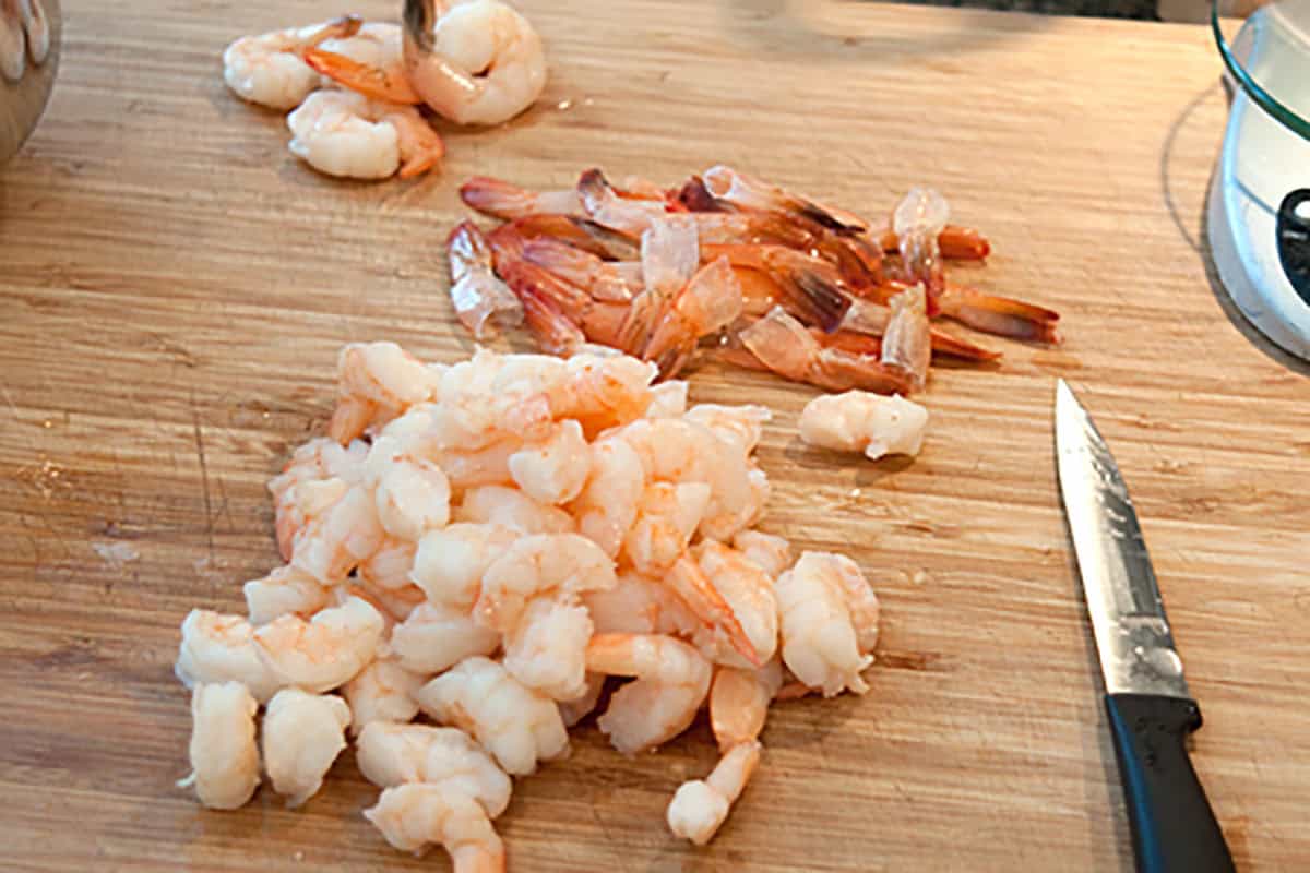Chopped shrimp and shrimp tails on a wooden cutting board with a knife nearby.
