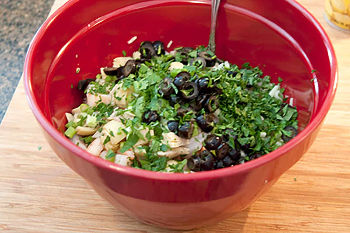 A red bowl filled with chopped vegetables, herbs, and sliced black olives, with a spoon inside.