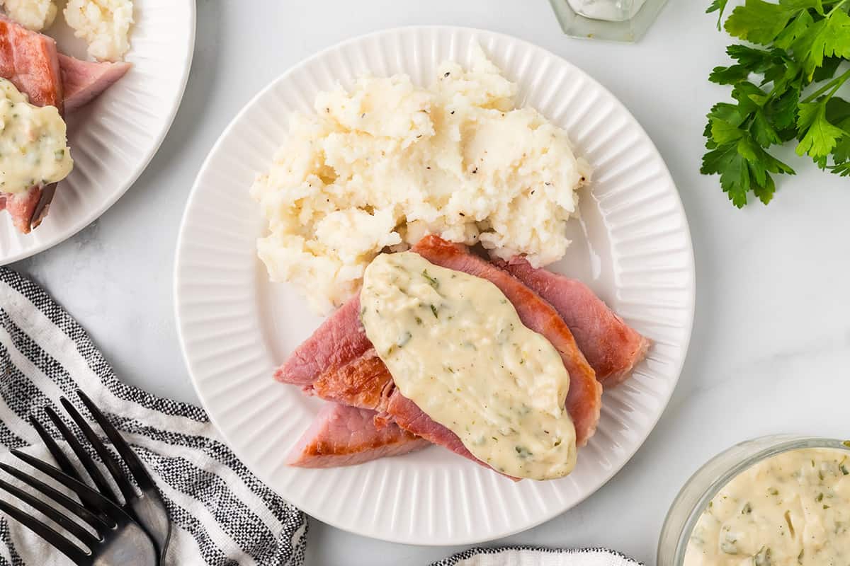A slice of ham topped with creamy sauce on a white dinner plate.