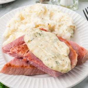 A slice of ham topped with creamy sauce on a white dinner plate.