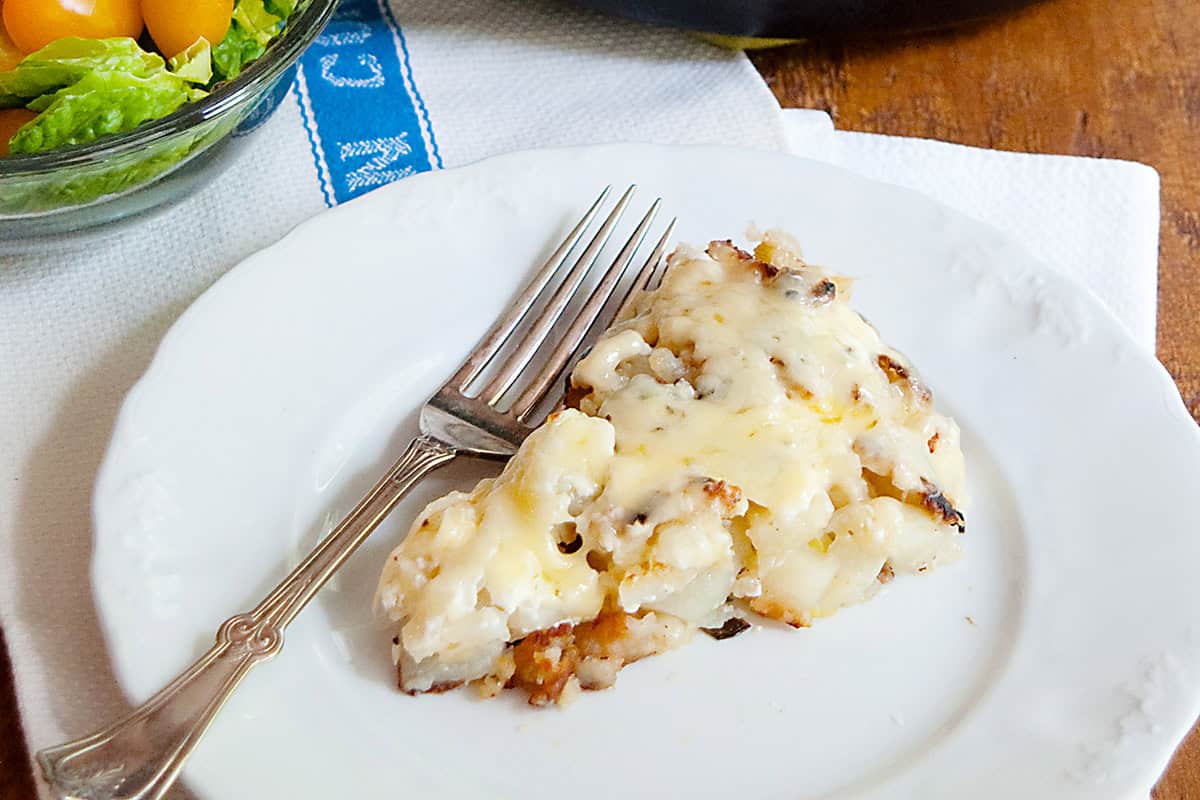 A slice of cheesy potato casserole on a white plate with a fork, next to a bowl of salad.
