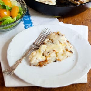 A slice of hash browns with gruyere on a white plate with a fork, next to a salad with tomatoes.