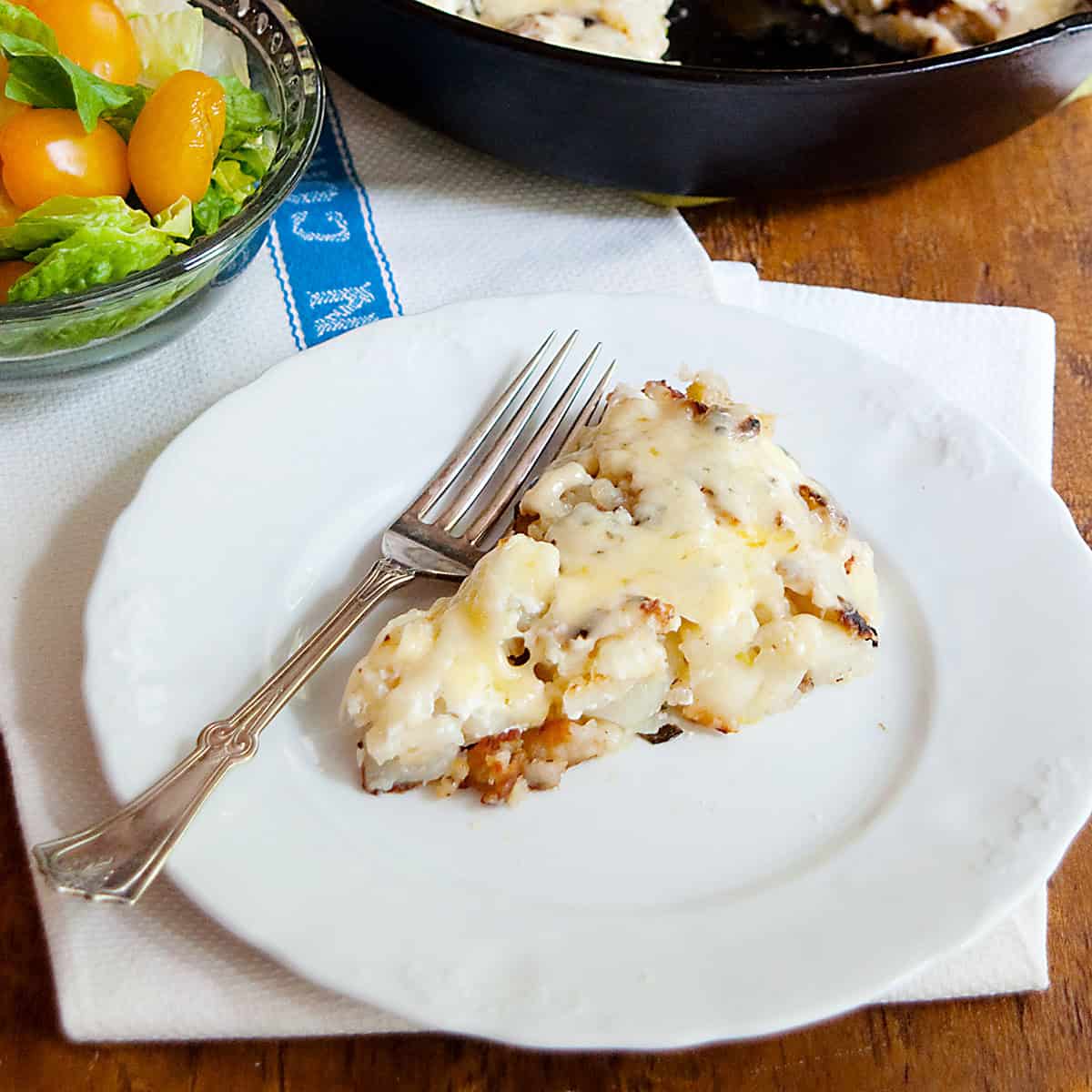 A slice of hash browns with gruyere on a white plate with a fork, next to a salad with tomatoes.