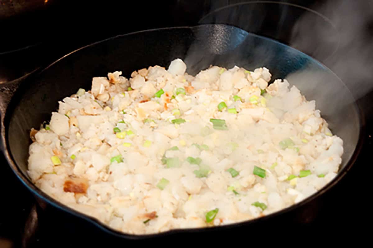Diced potatoes and green onions cooking in a steaming cast-iron skillet.