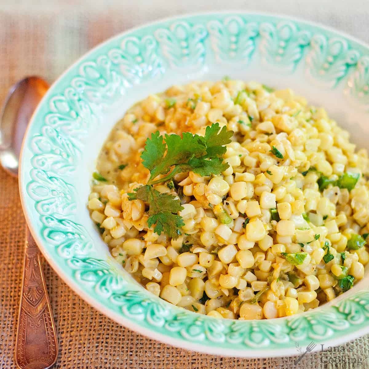 A plate of seasoned corn kernels garnished with fresh cilantro, served in a decorative bowl next to a spoon on a burlap tablecloth.