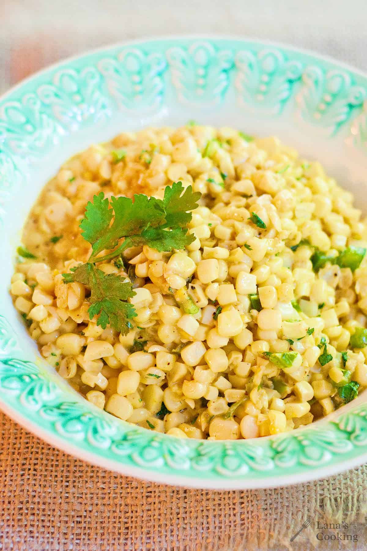 A bowl of creamy corn salad garnished with cilantro, served in a decorative green and white dish on a burlap surface.