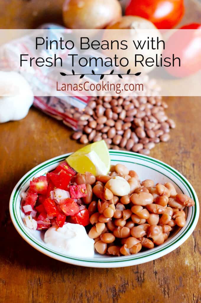 A bowl of cooked pinto beans with tomato relish, sour cream, and lime wedges on the side.