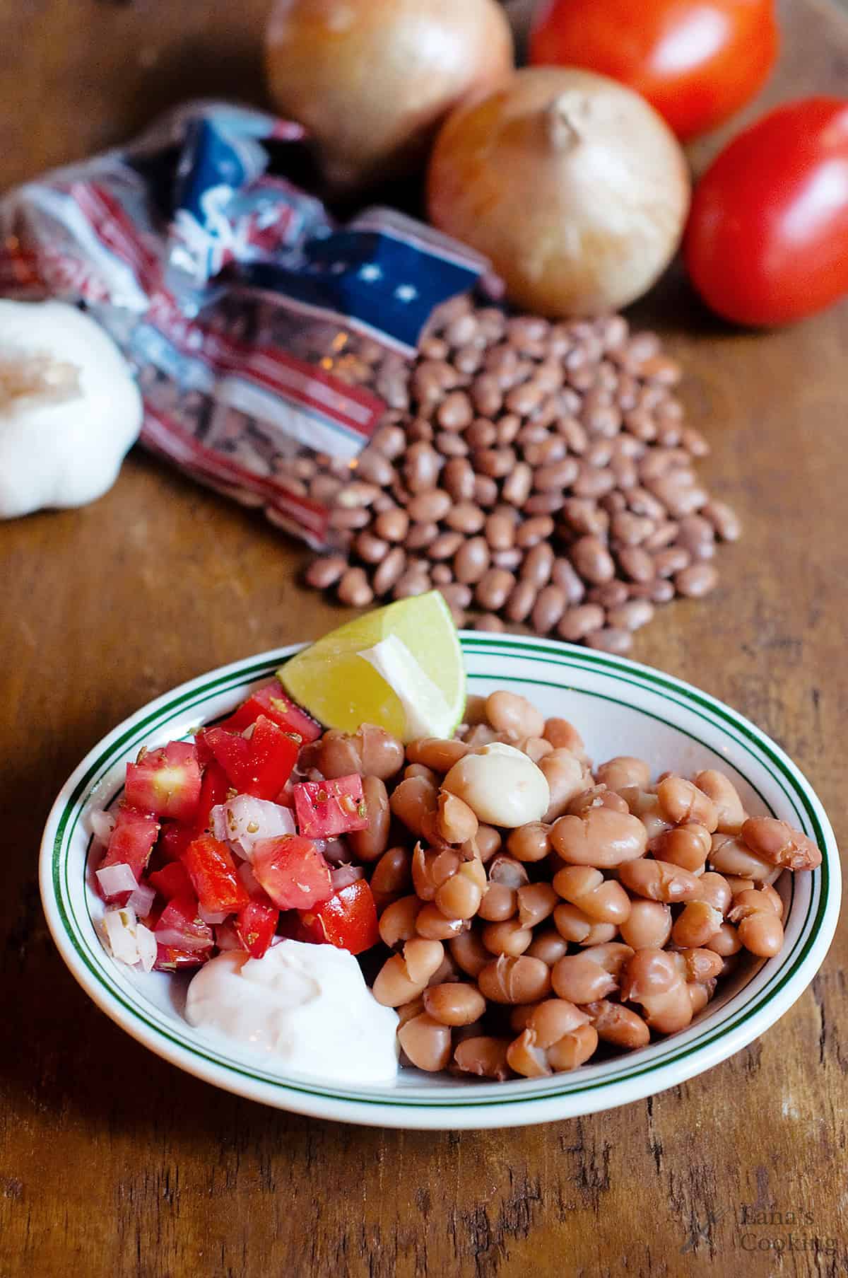 A plate with cooked pinto beans, a scoop of salsa, a lime wedge, a dollop of cream, and a clove of garlic. Raw beans, onions, tomatoes, and garlic are in the background.
