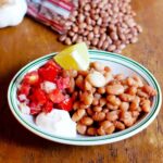 A bowl of cooked pinto beans with tomato relish, sour cream, and lime wedges on the side.