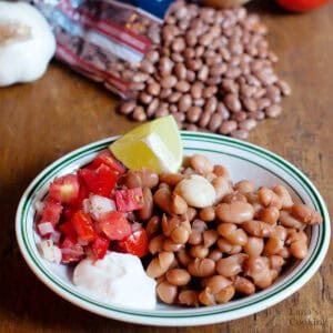 A bowl of cooked pinto beans with a garlic clove, diced tomatoes and onions, a lime wedge, and a dollop of sour cream. A bag of dried beans and garlic are in the background.