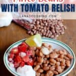 A plate with pinto beans, tomato relish, a lime wedge, garlic clove, and a dollop of sour cream sits on a table with dry beans and vegetables in the background.