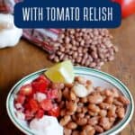 A plate with pinto beans, tomato relish, a lime wedge, and a dollop of sour cream, with raw beans, tomatoes, and garlic in the background.