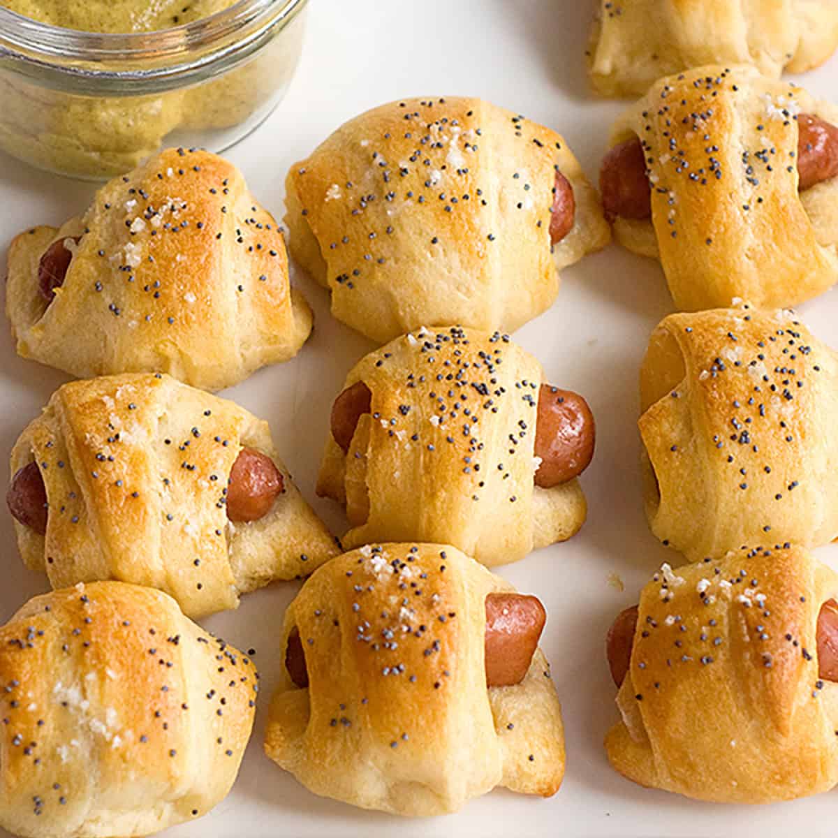Sausage and pineapple rollups topped with poppy seeds and salt, arranged on a white plate with mustard on the side.