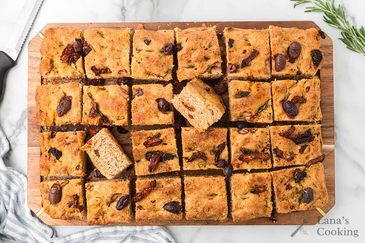 Sliced focaccia bread with olives and sun-dried tomatoes on a wooden board, viewed from above.