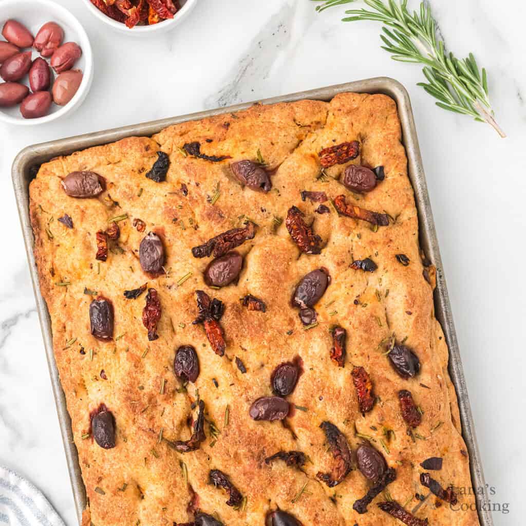 Golden focaccia bread topped with Kalamata olives, sun-dried tomatoes, and rosemary on a baking tray.