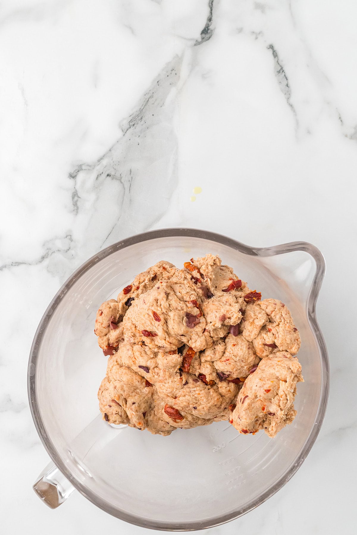 Brown dough with visible pieces of dried fruit in a clear mixing bowl on a white marble surface.