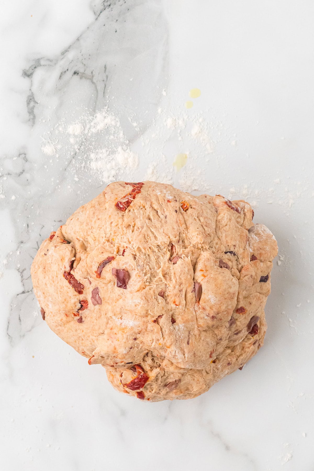 A ball of rustic bread dough with sun-dried tomatoes on a floured marble surface.