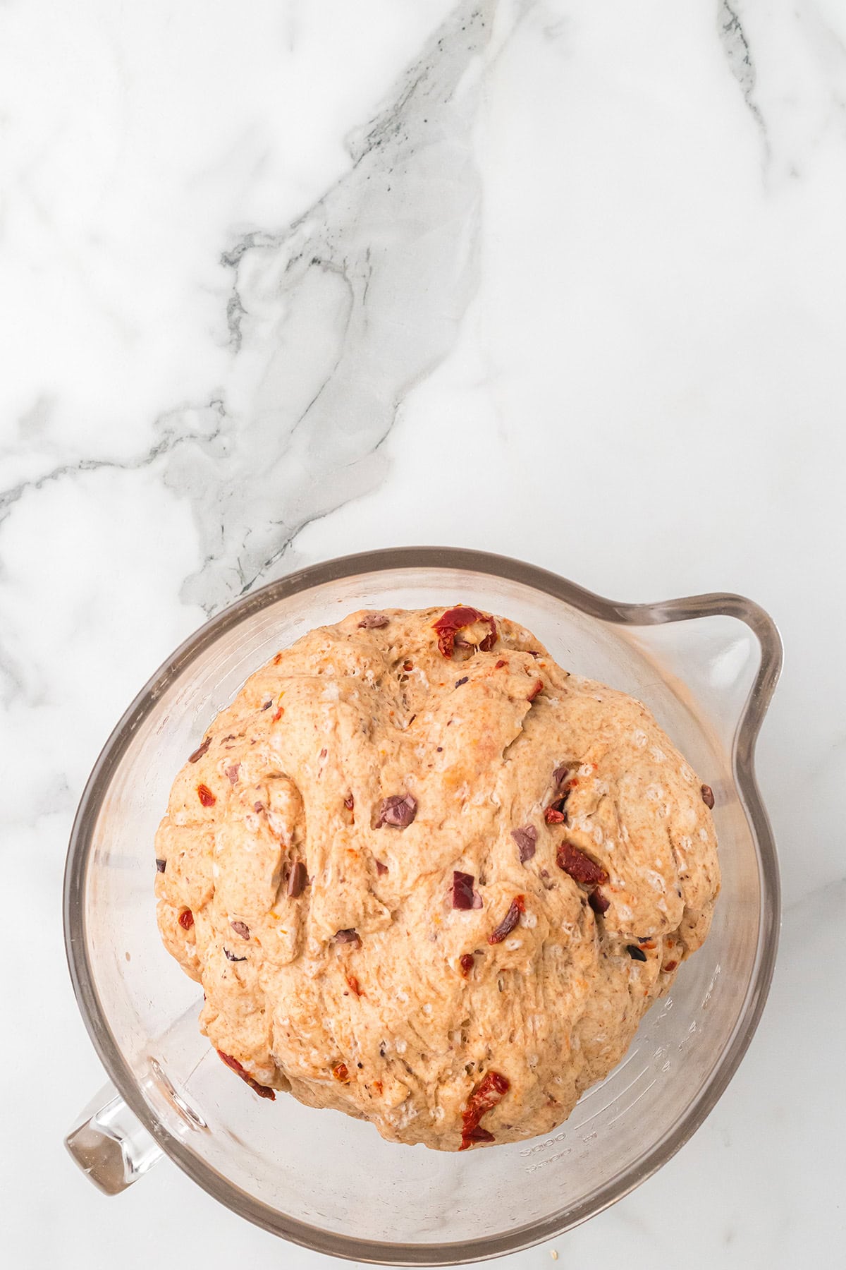 A glass bowl of dough with visible pieces of sun-dried tomatoes on a white marble surface.
