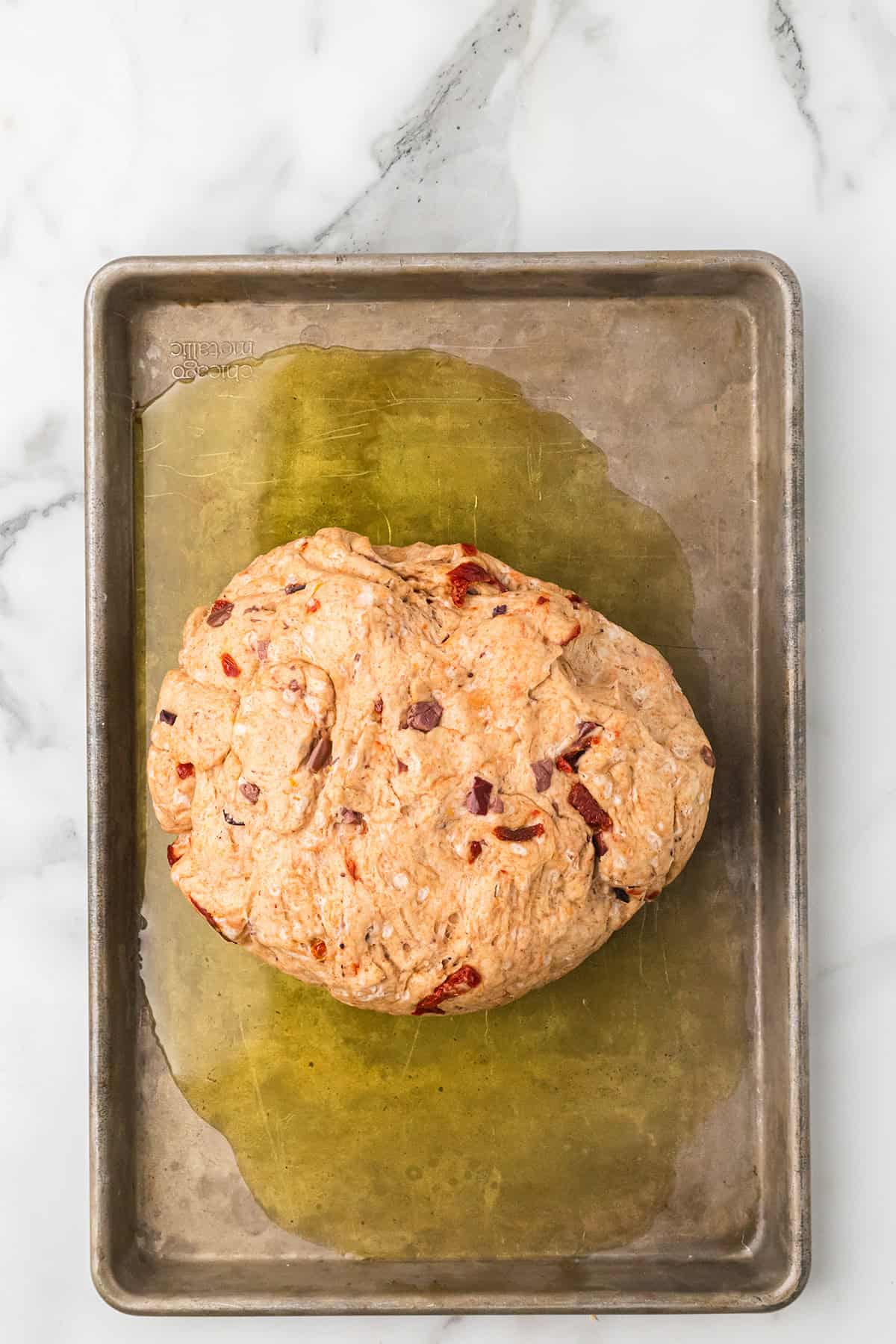 A ball of dough with sun-dried tomatoes on an oiled baking sheet, set on a marble surface.