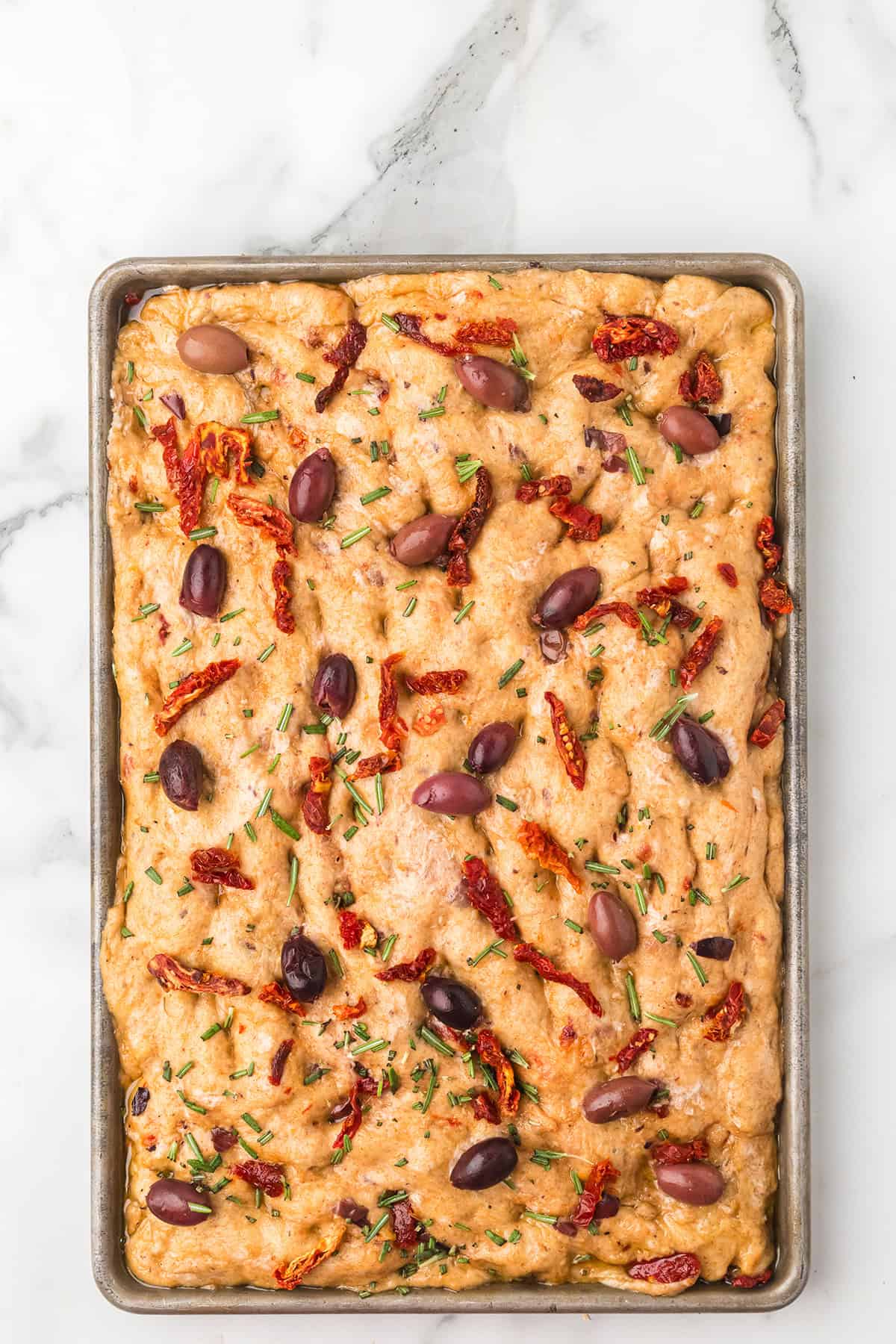 A rectangular focaccia topped with olives, sun-dried tomatoes, and rosemary on a marble surface.
