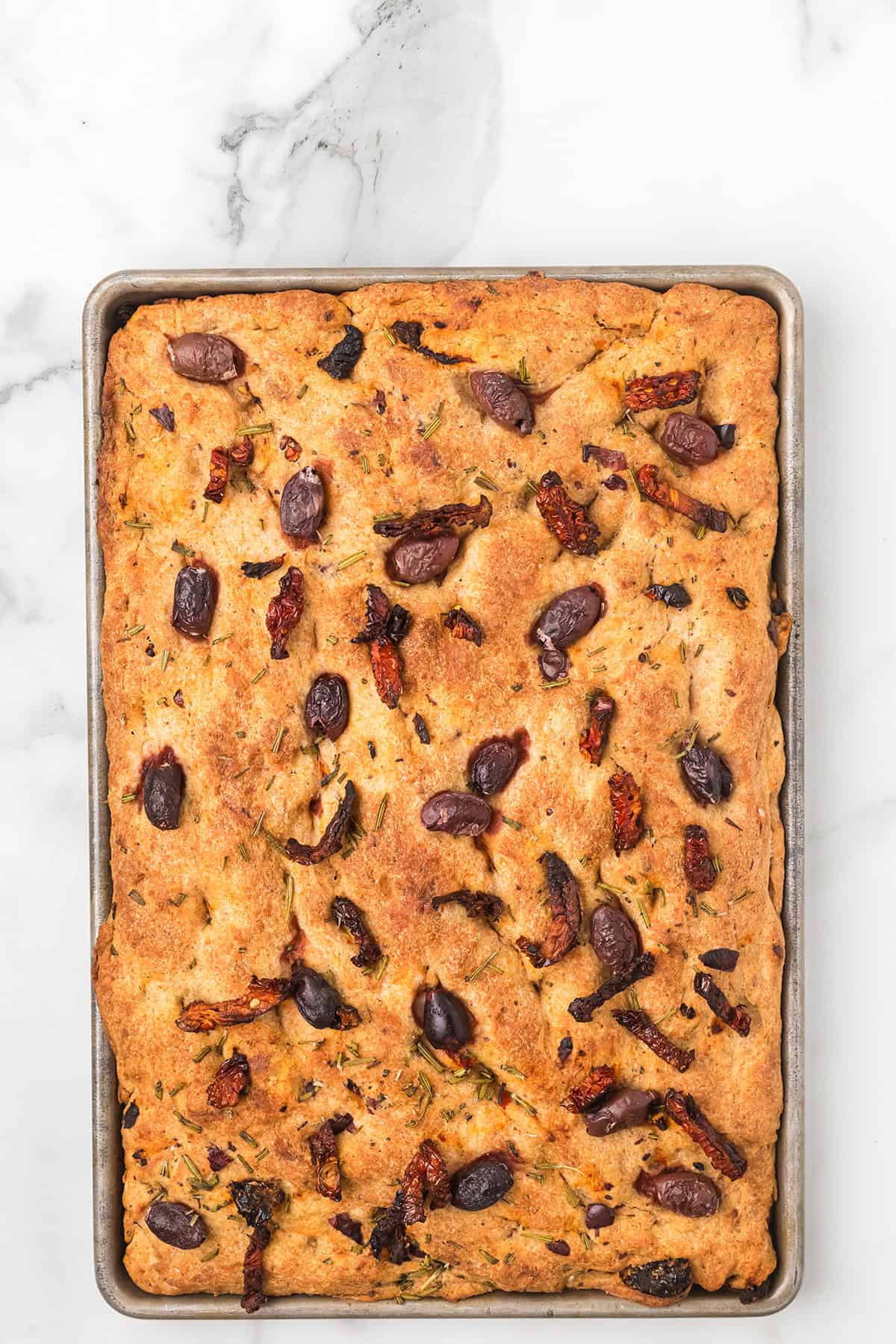 A baked sheet of focaccia bread topped with olives, herbs, and sun-dried tomatoes on a marble surface.