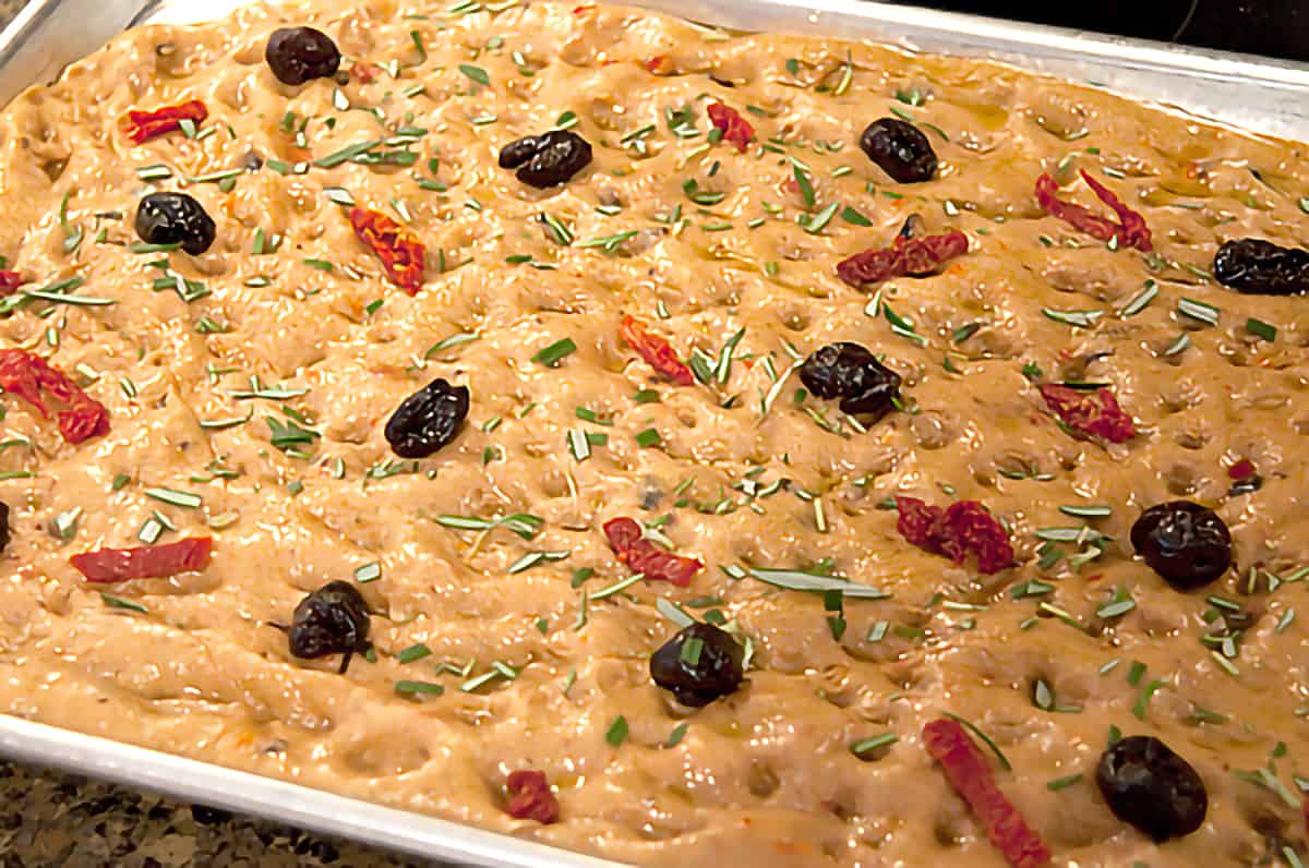 Finished focaccia dough in a baking pan with additional olives and tomatoes on top sprinkled with chopped fresh rosemary.