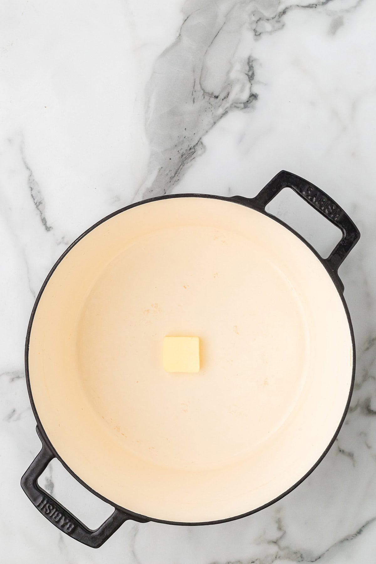 A single cube of butter sits in the center of an empty cream-colored Dutch oven on a marble surface.