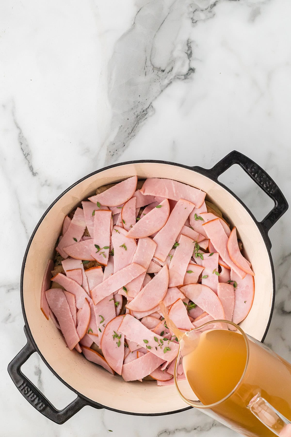 Ingredients for Dublin Coddle in a pot, as broth is being poured over them on a marble countertop.