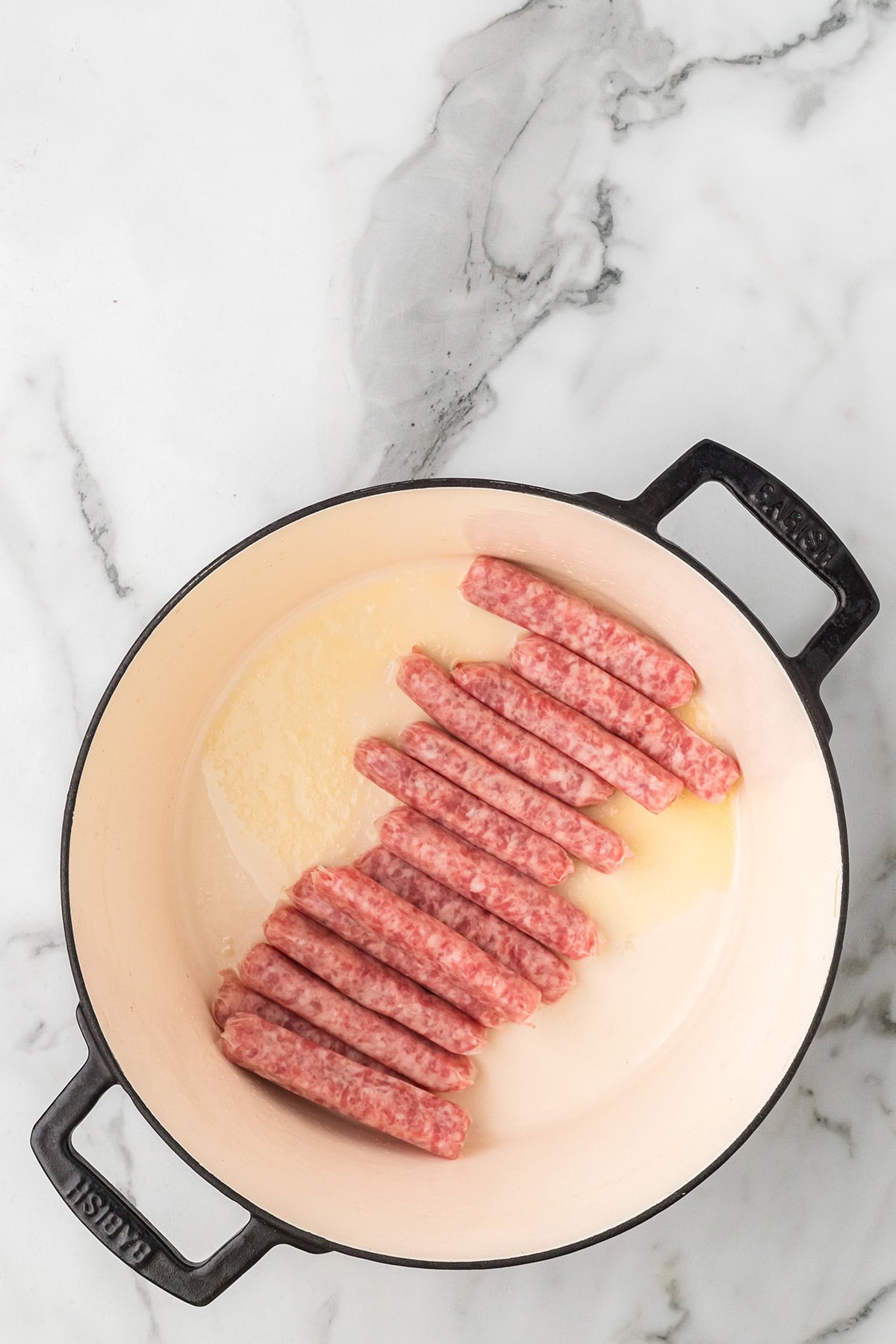 Raw sausage links arranged in a white Dutch oven on a marble countertop.
